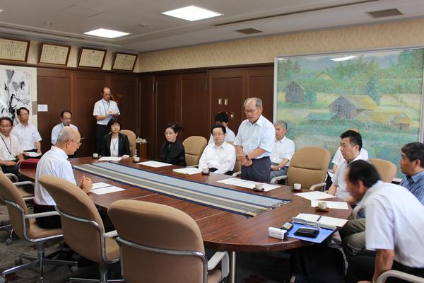 向かいに座る市長を前に、立っているご年配の男性とその周りに座っている関係者の男女の写真