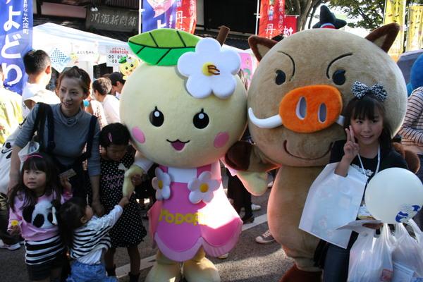 まるいのの着ぐるみと頭にお花を付けた着ぐるみが並んでいて近くに親子連れの人や子供がピースして一緒に記念写真をとっている写真