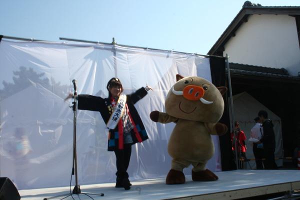 まるいのの着ぐるみと観光大使と書かれたタスキをかけた女性が舞台に立っている写真