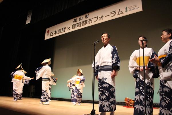 舞台の上で、浴衣のような衣装をきて鳥追い傘を被った人が、横の方で歌っている歌に合わせて踊っている写真