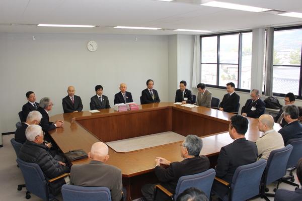 四角になったテーブルにスーツ姿の20人ほどの男性が座って会議している写真