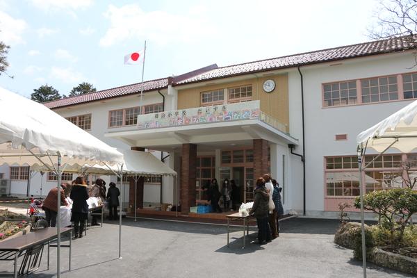 木造の校舎の入り口の前で女性たちが机に袋を並べて立っている写真