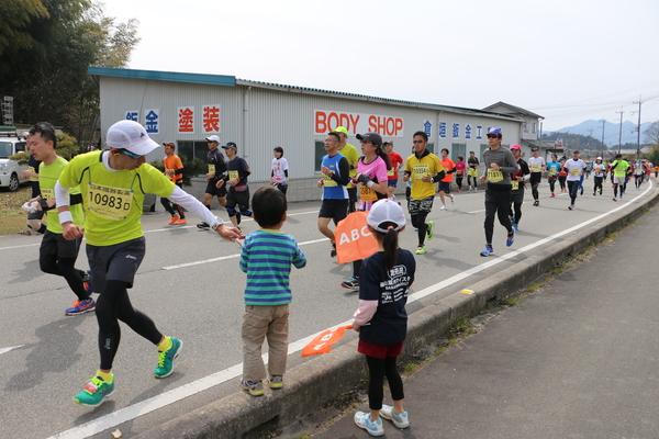 沿道でABCの旗を持った小さな男の子と女の子がランナー達を応援している写真