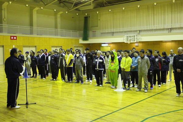 体育館に市の職員が運動しやすいジャージを身に着け、整列して並んでおり、その前で男性がスタンドマイクで話をしている様子の写真
