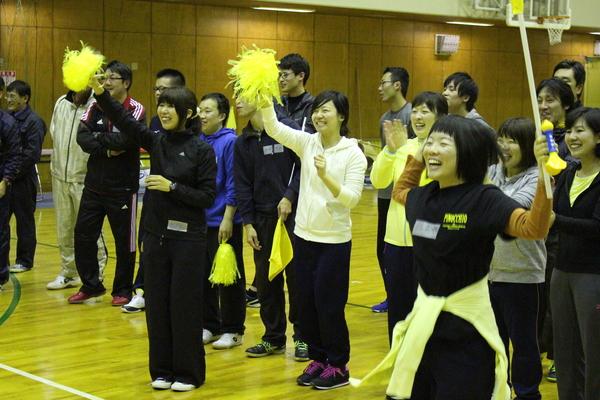 競技に参加していない職員の方々が黄色いポンポンをもって立ちあがって応援しており、応援席でも盛り上がっている様子の写真