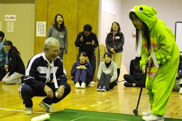 グリーンのコートが敷かれて囲碁ボールが行われており、ボールを持った白髪の男性しゃがんで笑っており、その横にはガチャピンの衣装を着た女性がスティックを握ってい構えている様子の写真