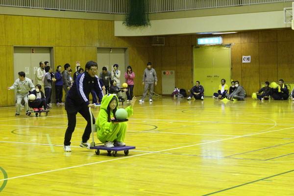 2人1組で、男性が女性を台車に乗せて押しており、台車の女性はお盆にボールを載せてバランスを取って乗っている様子の競技を写している写真