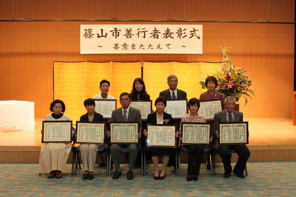 篠山市善行者表彰式と書かれている横断幕と金屏風の前で、受賞者9名が額に入った表彰状を持って写っている記念写真