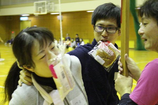 パン食い競争をしており、男性がアンパンを食わえており、女性がパンを食わえようとしている様子の写真
