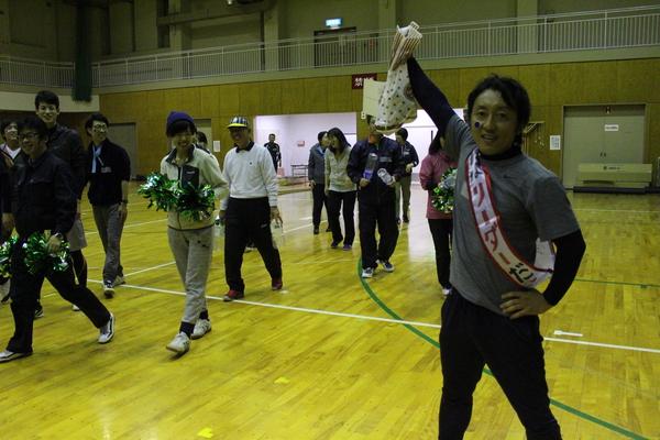 肩に「リーダー」と書かれているタスキをかけている男性が右手に袋を持って上に挙げており、その様子を見ている職員の方々が歩きなら笑っている様子の写真
