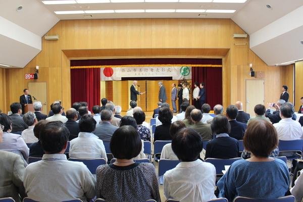 舞台の金屏風の前で、市長より受賞者が賞状を読んでもらっている様子を、観客席に座っている多くの人が見ている様子の写真