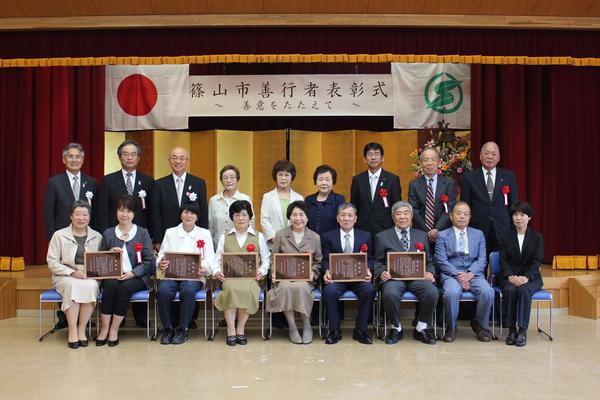篠山市善幸者表彰式と書かれている横断幕と金屏風の舞台の前で、受賞者が額に入った賞状を手に持って笑顔で写っている記念写真
