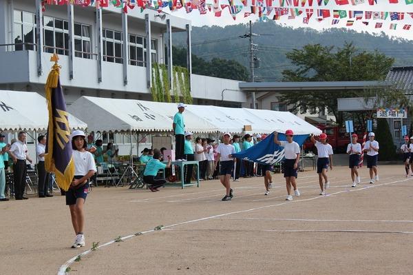 校旗を持っている女生徒の後ろに4名の生徒が幕を持ち、その後方にはプラカードを持った生徒がグラウンドを行進している写真