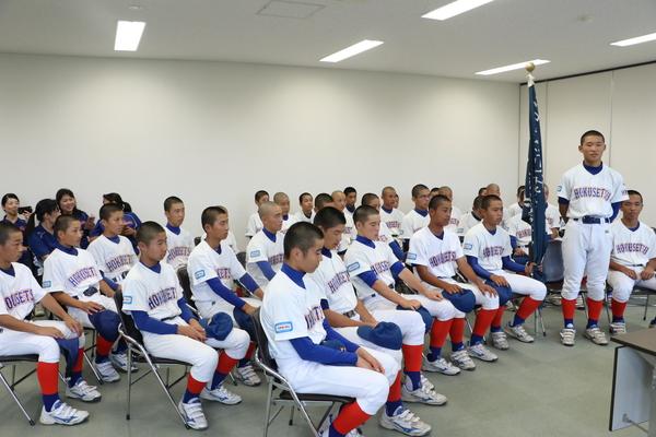 野球部が並んで座り、1名の代表選手が後ろに手を組み立ち話をしている写真