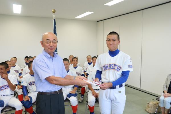 市長から奨励金を受け取る男子選手の後ろにユニフォームを着て座る野球部員の写真