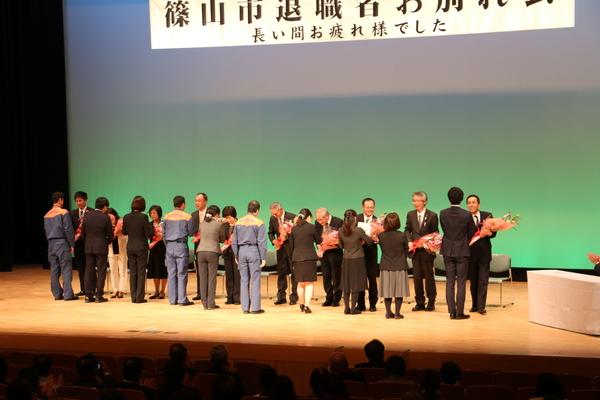 ステージの上で関係者たちが退職者全員に花束を同時に渡している写真