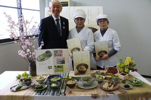 郷土料理のレシピ本「よろしゅうおあがり」が完成し講師の2名が白のかっぽう着と調理用の帽子を被り両手に本を持ち市長が左に立ち目の前にレシピ本通りに作られた料理の数々を目の前に置いて記念撮影の写真