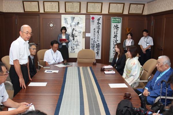 楕円形のテーブルが置いてある椅子に6名ほどの男女が座っていて、市長が立ち上がって話をしている写真