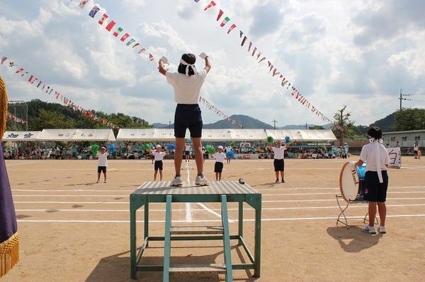 ハチマキをした生徒が台の上で両手を上げて立ち、グラウンドに立っている生徒と向かい合い、その横にはハチマキをして太鼓を叩いている生徒が写っている写真