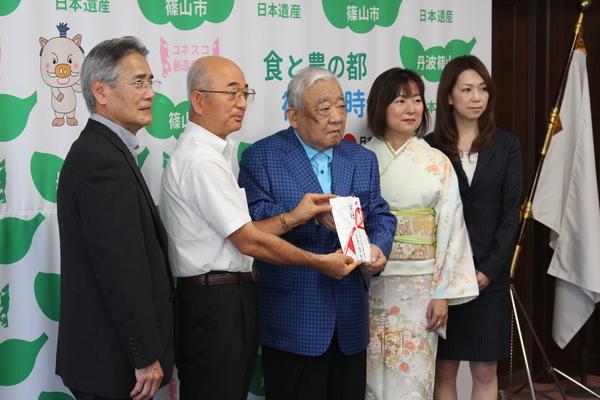 市長と男性が祝儀袋を二人で持ちその横にスーツを着た男性一名、女性一名、着物を着た女性が一名並んでいる写真