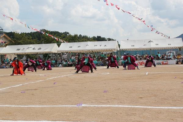 グラウンド内にて赤の長い法被を着て腰を落としながら踊っている人々の写真