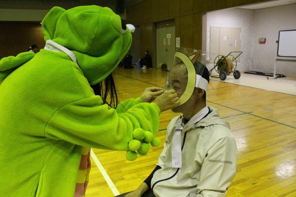 ガチャピンの着ぐるみ姿の女性が男性にお面をかぶせている写真