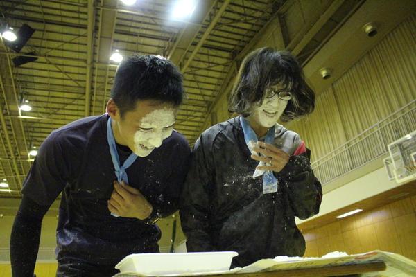 飴食い競争にて、男性は笑顔で真っ白の顔して、女性は眼鏡も顔も真っ白になった写真