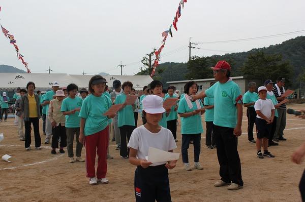 生徒の後ろで緑色のTシャツを着た方々が手に持っている紙を見て声に出し、赤の帽子を被った男性が女性にマイクを向けている写真