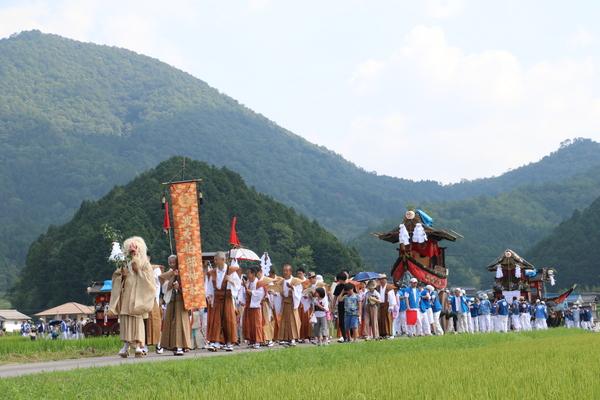 田んぼ道をお面を被り玉串を持った人を先頭に数基の神輿を担いで練り歩く人々の写真