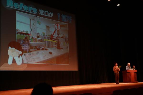 チームで取り組んだ改善案をスクリーンで男性が発表している写真