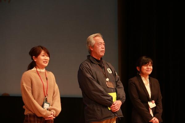 女性職員2名と男性職員1名が壇上で両手を前に添えて立っている写真