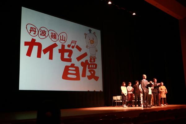 「丹波篠山カイゼン自慢」と映し出されたスクリーンの前に約10名の人が横2列に並んで立っている写真