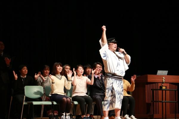 壇上でマイクを持ち学生帽を被り浴衣を着た男性が右手で握り拳を作りうえにあげ、その後ろで椅子に座った女性や男性が手拍子をしている写真