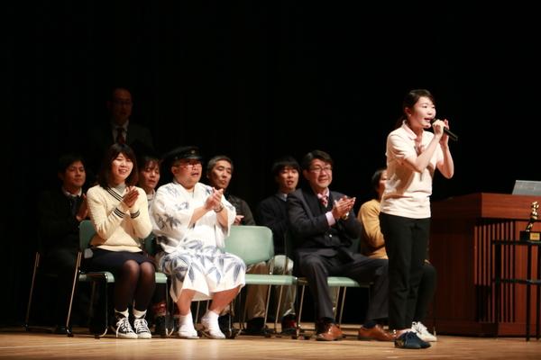 女性の方が両手でマイクを持ち歌い、その後ろで椅子に座り手拍子をしている男性や女性の写真