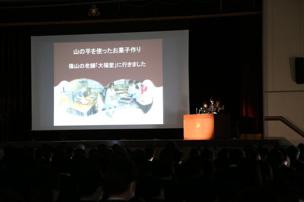 照明が落ちた暗い中、舞台の上で山の芋を使ったお菓子作りという紹介スライドの横で3人の高校生が話をしている写真