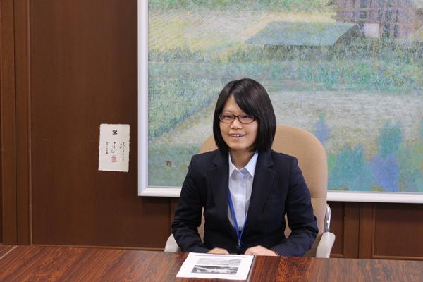 テーブルに資料が置いてあり、酒井克美さんが椅子に座り挨拶をしている写真