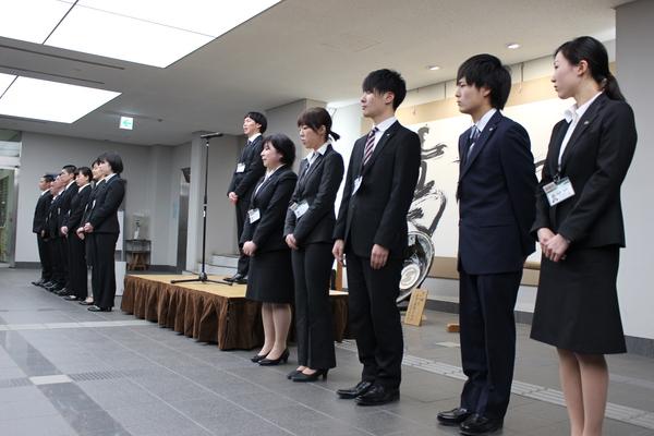新規職員が横1列に並び、そのうち1名の男性職員がマイクの前で話しをしている写真