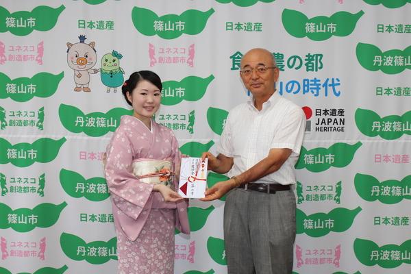 篠山市と書かれた壁の前で小さい花の模様が入った着物を来た小林さんが市長から奨励金を受け取っている写真
