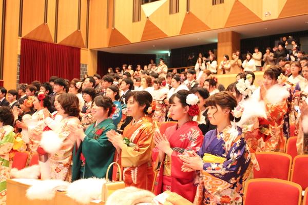 晴れ着姿やスーツ姿の新成人者が成人式の客席に立って拍手をしている写真