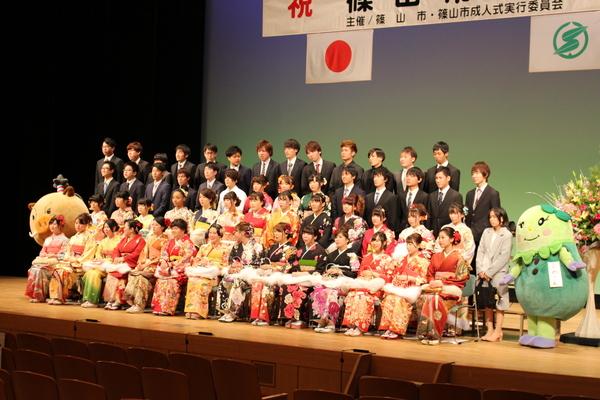 舞台で晴れ着姿やスーツ姿の新成人者がゆるキャラと一緒に記念撮影している様子を右斜め前から写した写真