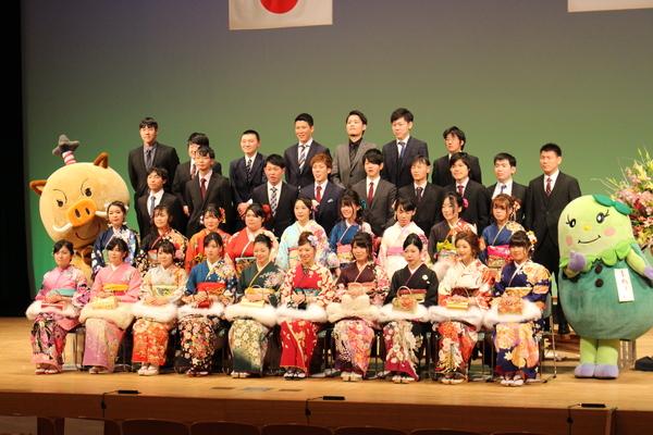 舞台で晴れ着姿の女性成人者やスーツ姿の男性成人者がゆるキャラと一緒に正面で記念撮影している写真