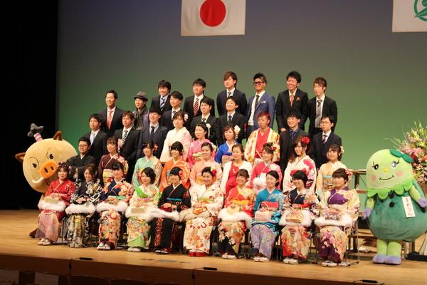 舞台で晴れ着姿の女性成人者や袴姿やスーツ姿の男性成人者がゆるキャラと一緒に少し斜めから記念撮影している写真