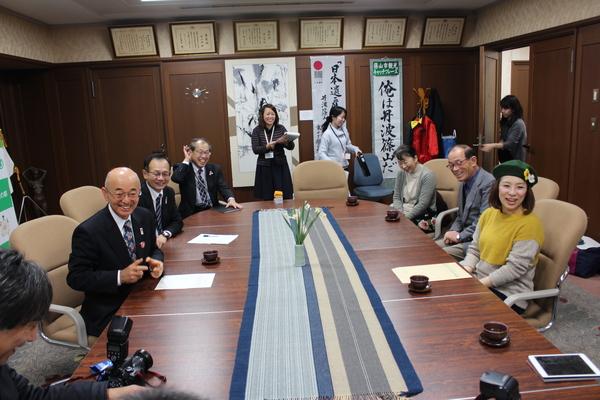 市長室にて、緑色に黄色と白の花が飾られているベレー帽をかぶった森田 まりこさんが市長の前に座っており、市長室にいる関係者が全員笑顔になっている様子の写真