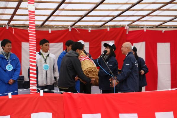 市長が紅白幕が張られたステージで男性に俵を手渡し、その様子を見ている男性の方々の写真