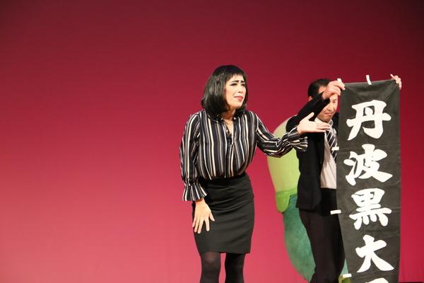 舞台でスーツ姿の男性が持っているのぼり旗の方に手で案内をしているストライプシャツとタイトスカート姿の女性の写真