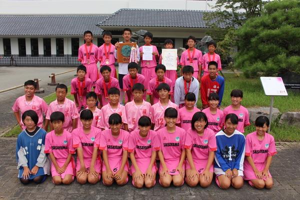 丹南中学校ホッケー部の生徒(前列で笑顔で写っている女生徒、後列でメダルを首に下げたり、賞状や盾を手に持っている男子生徒)の集合写真
