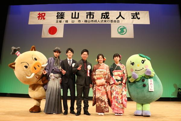 舞台にて袴姿やスーツ姿の男性成人者と晴れ着姿の女性成人者の両脇にゆるキャラが一緒に並んで記念撮影している写真