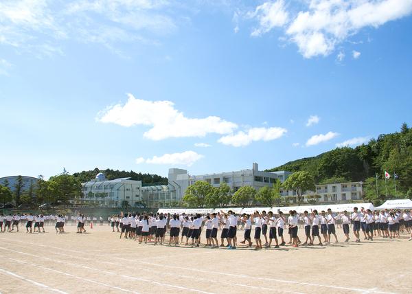 運動場で、生徒たちが輪になり手と足をあげて踊っている写真