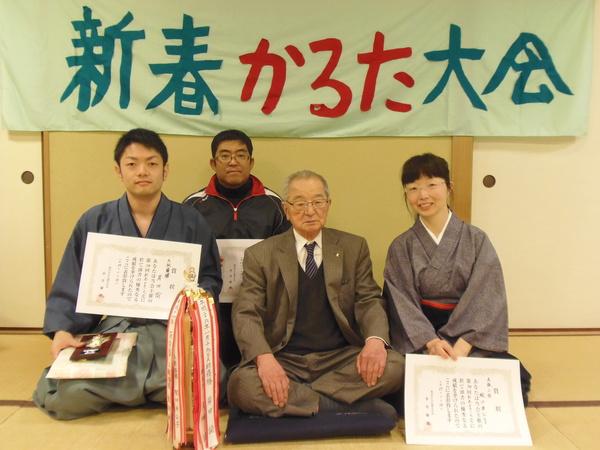 賞状をもって並ぶ男女と、座布団の上で胡坐をかく男性の集合写真