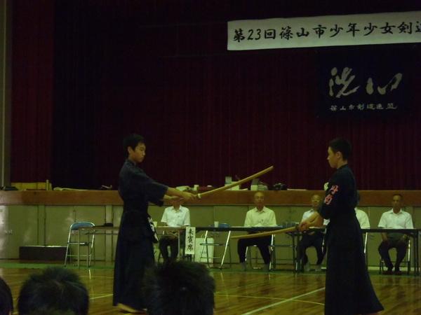 体育館の中で4名の男性が座っていて、その前で剣道着の姿の少年2名が竹刀を持って向かい合わせで立っている写真